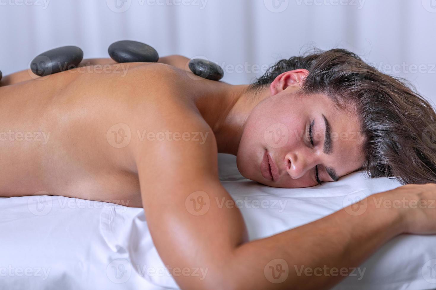 Close up view of beautiful young woman lying face down in a massage table with spa stones on her back photo