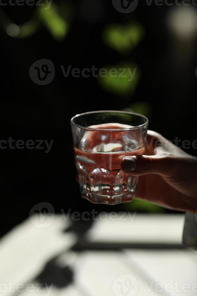 manos de mujer aisladas, sosteniendo un vaso de agua sobre un fondo oscuro con hojas verdes. los rayos del sol caen sobre el cristal. mañana saludable foto