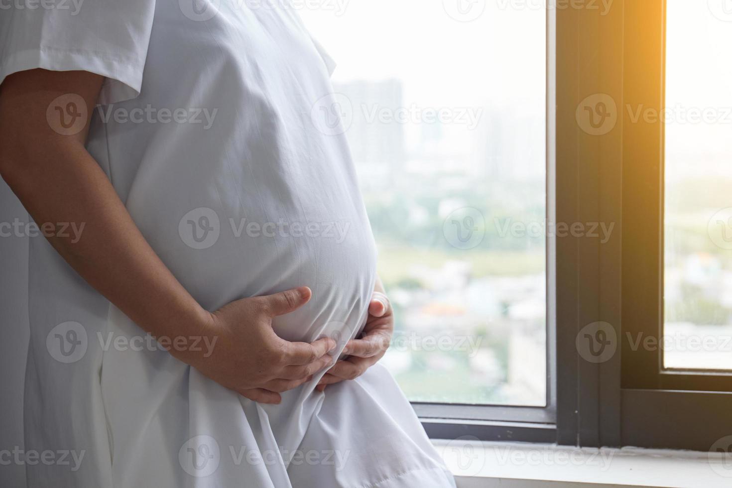 Photo of Happy Asian pregnant woman sitting in bed and touching her belly at home . With Small shoes for the unborn baby in the belly