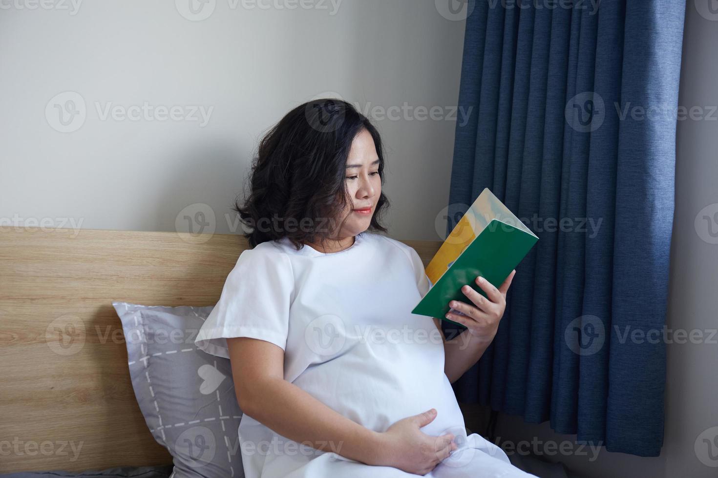Young Asian Pregnant Woman reading book on the bed at home while holding her belly photo