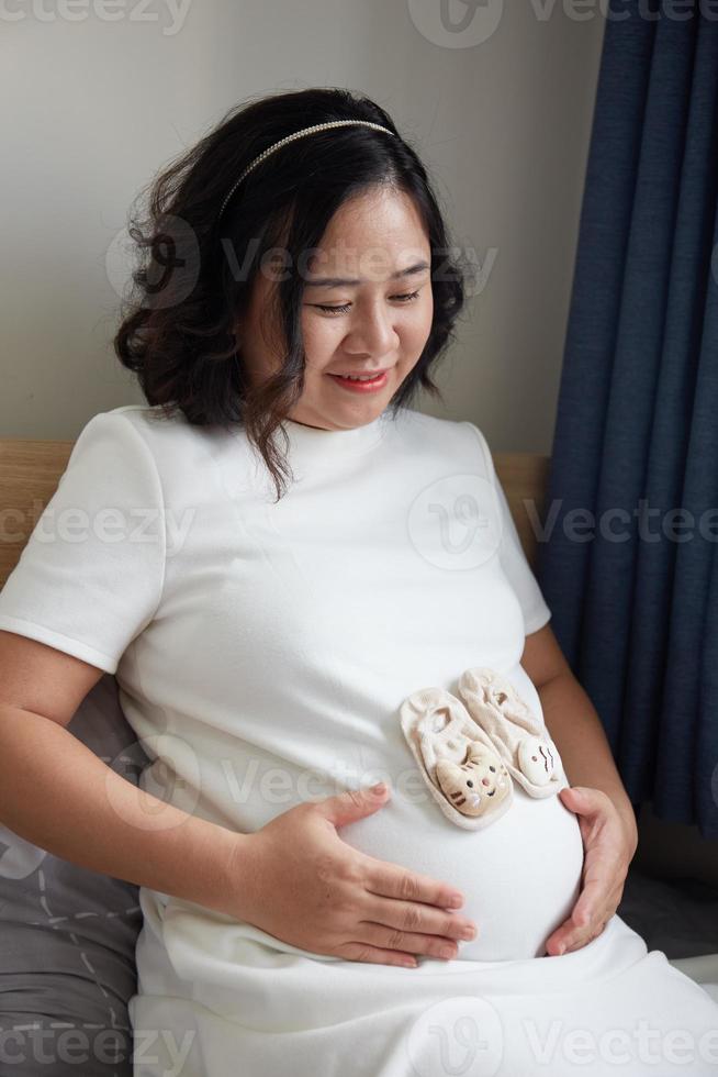 foto de contento asiático embarazada mujer sentado en cama y conmovedor su barriga a hogar . con pequeño Zapatos para el no nacido bebé en el barriga