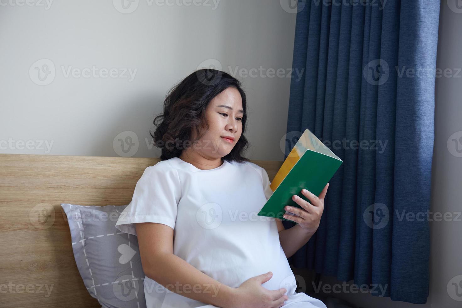 Young Asian Pregnant Woman reading book on the bed at home while holding her belly photo