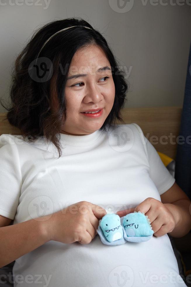 Photo of Happy Asian pregnant woman sitting in bed and touching her belly at home  With Small shoes for the unborn baby in the belly