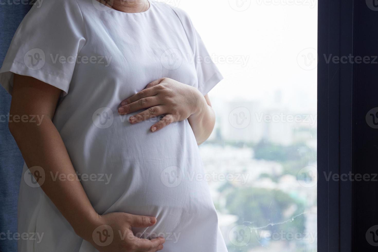 Photo of Happy Asian pregnant woman sitting in bed and touching her belly at home . With Small shoes for the unborn baby in the belly