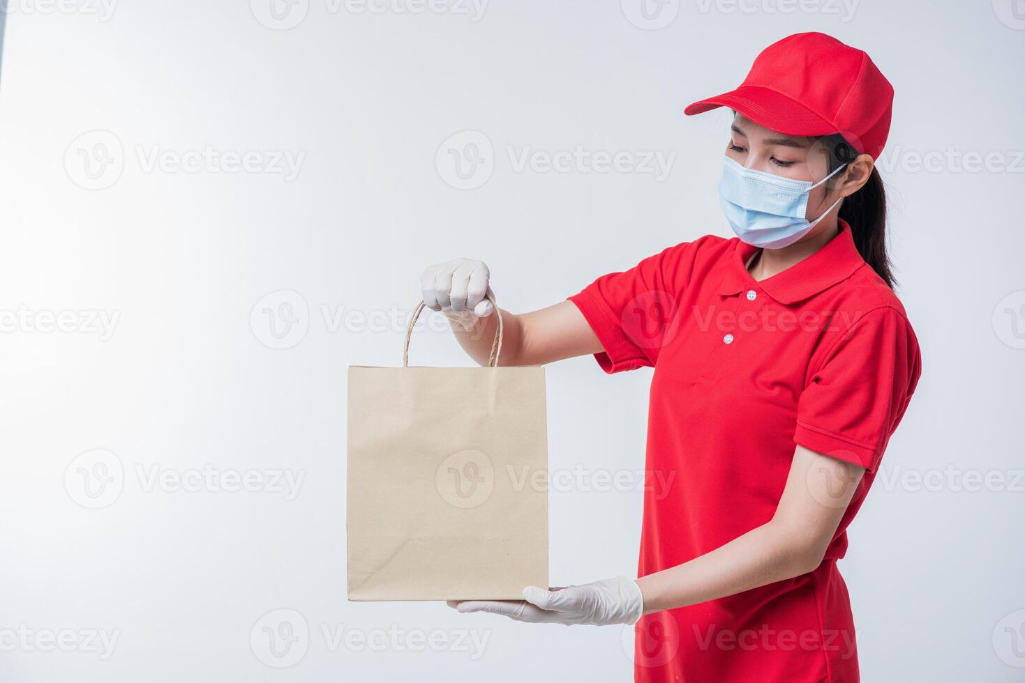 imagen de un joven repartidor feliz con gorra roja en blanco camiseta uniforme mascarilla guantes de pie con un paquete de papel artesanal marrón vacío aislado en un estudio de fondo gris claro foto