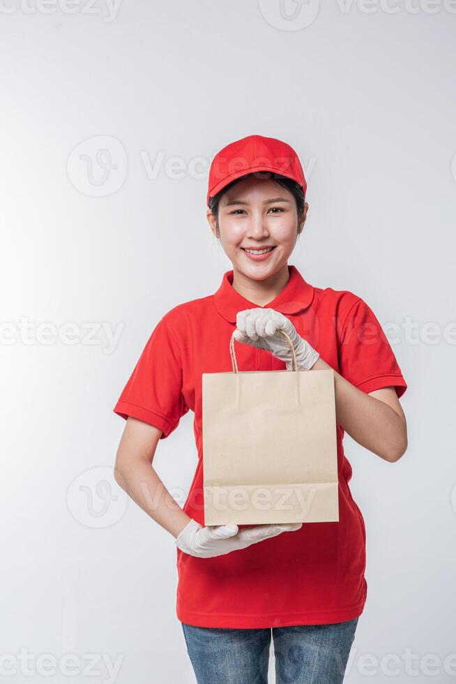 imagen de un joven repartidor feliz con gorra roja en blanco uniforme de camiseta de pie con un paquete de papel artesanal marrón vacío aislado en un estudio de fondo gris claro foto