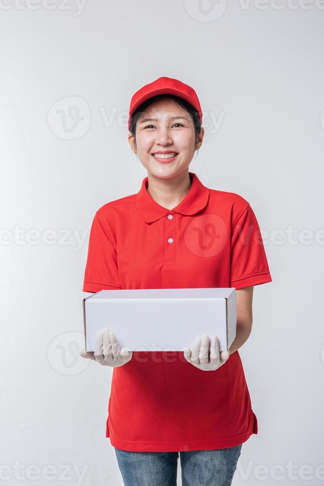imagen de un joven repartidor con uniforme de camiseta en blanco con gorra roja de pie con una caja de cartón blanca vacía aislada en un estudio de fondo gris claro foto