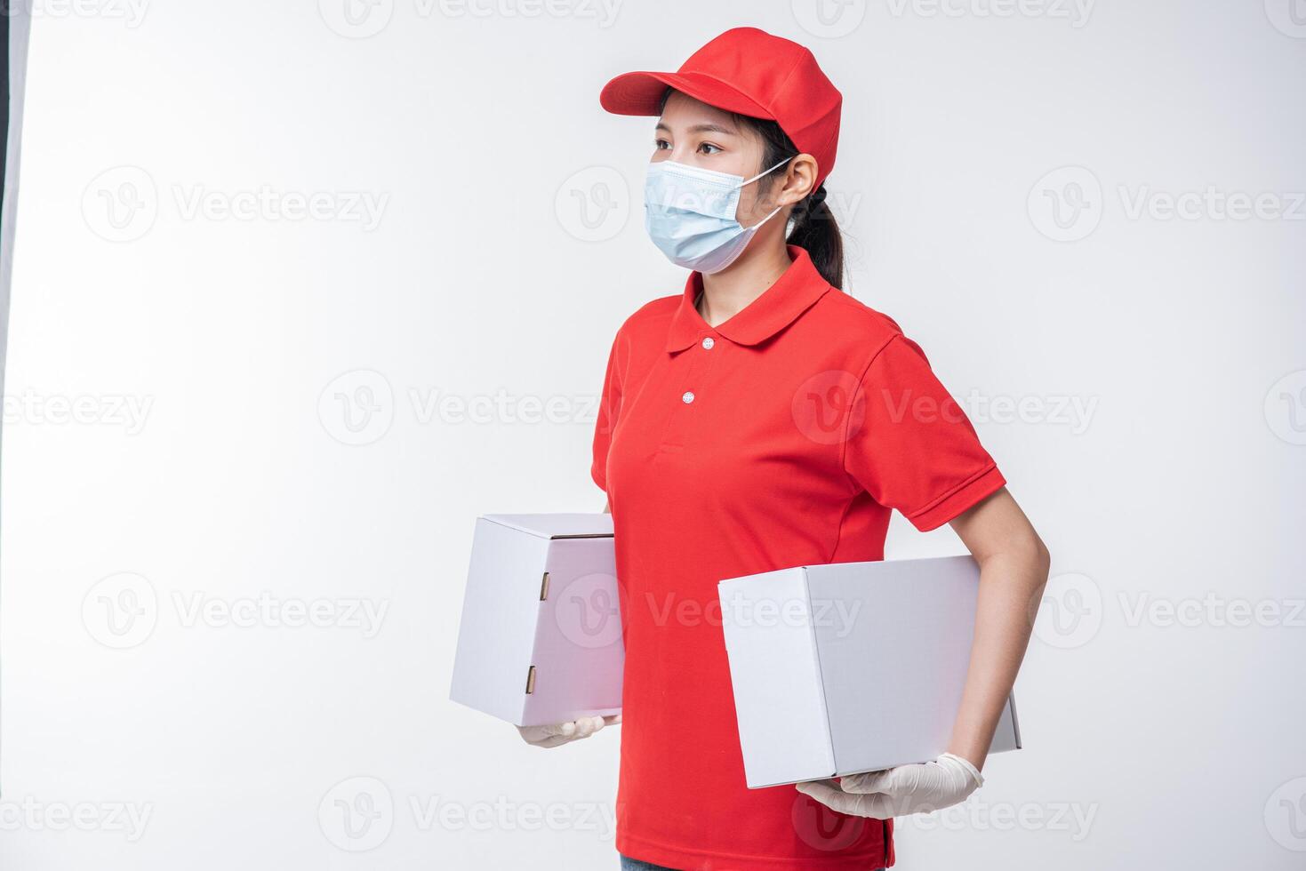 Image of a conscious young delivery man in red cap blank t-shirt uniform face mask gloves standing with empty white cardboard box isolated on light gray background studio photo