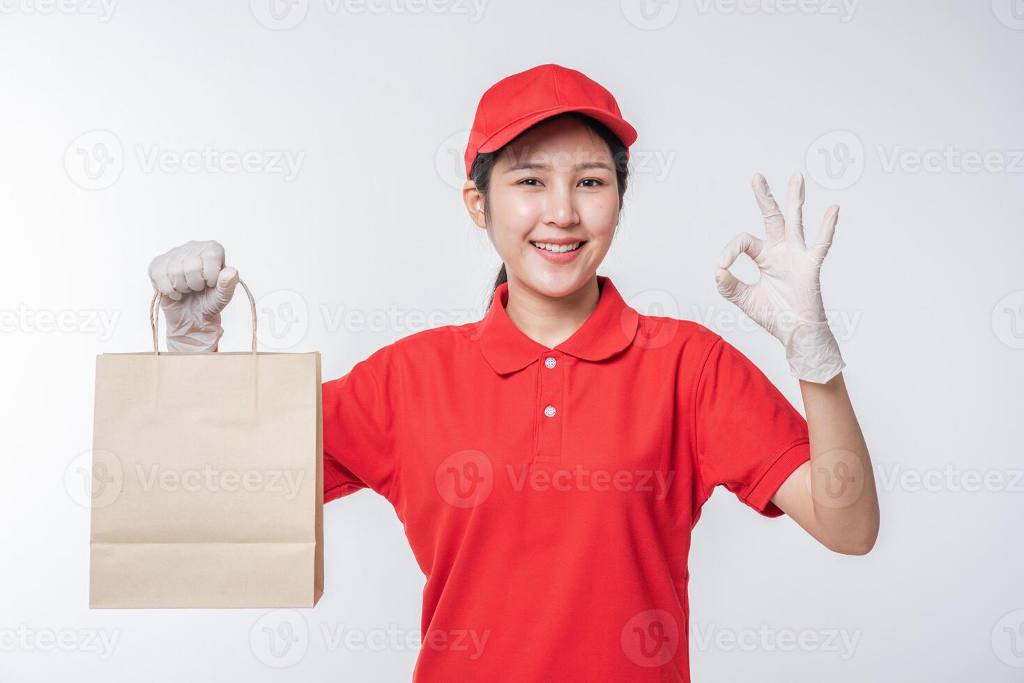 Image of a happy young delivery man in red cap blank t-shirt uniform standing with empty brown craft paper packet isolated on light gray background studio photo