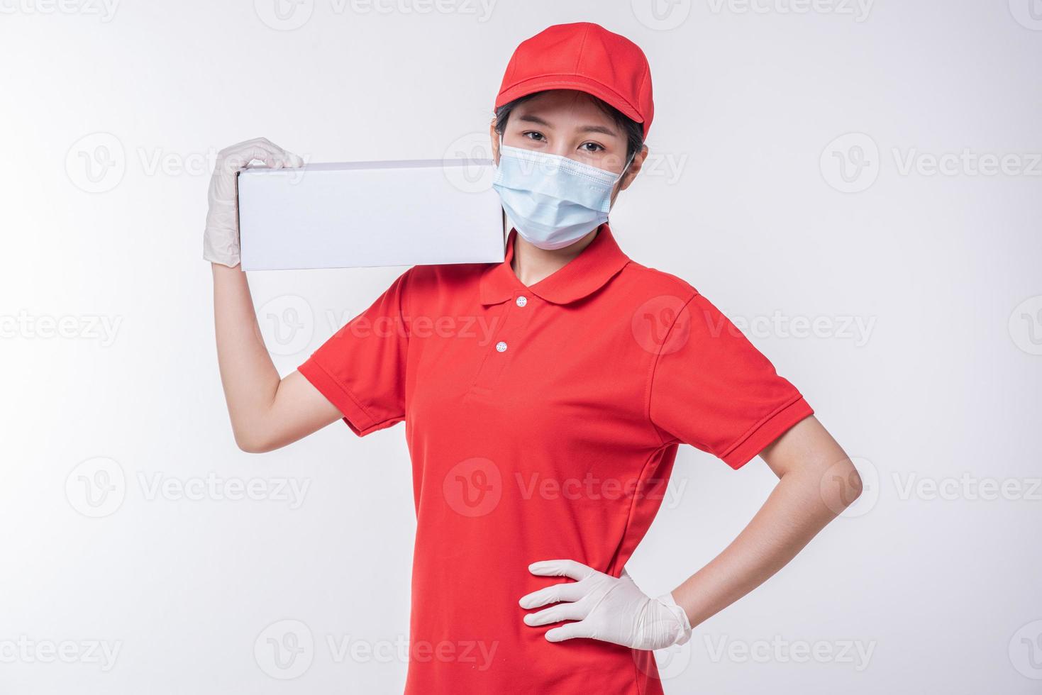 imagen de un joven repartidor consciente con gorra roja en blanco, camiseta uniforme, guantes de máscara facial de pie con una caja de cartón blanca vacía aislada en un estudio de fondo gris claro foto