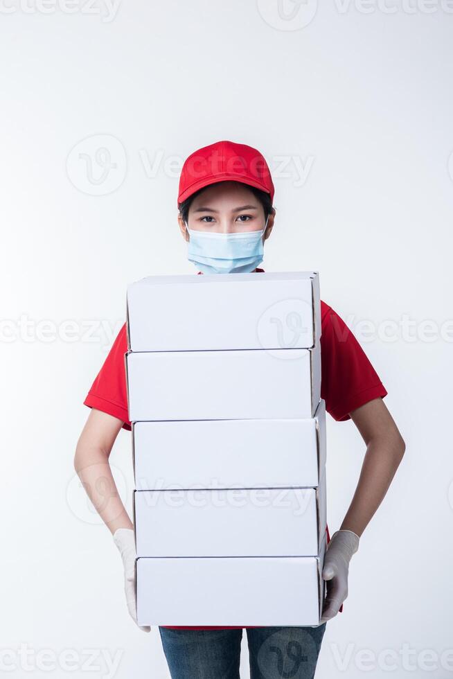 imagen de un joven repartidor consciente con gorra roja en blanco, camiseta uniforme, guantes de máscara facial de pie con una caja de cartón blanca vacía aislada en un estudio de fondo gris claro foto