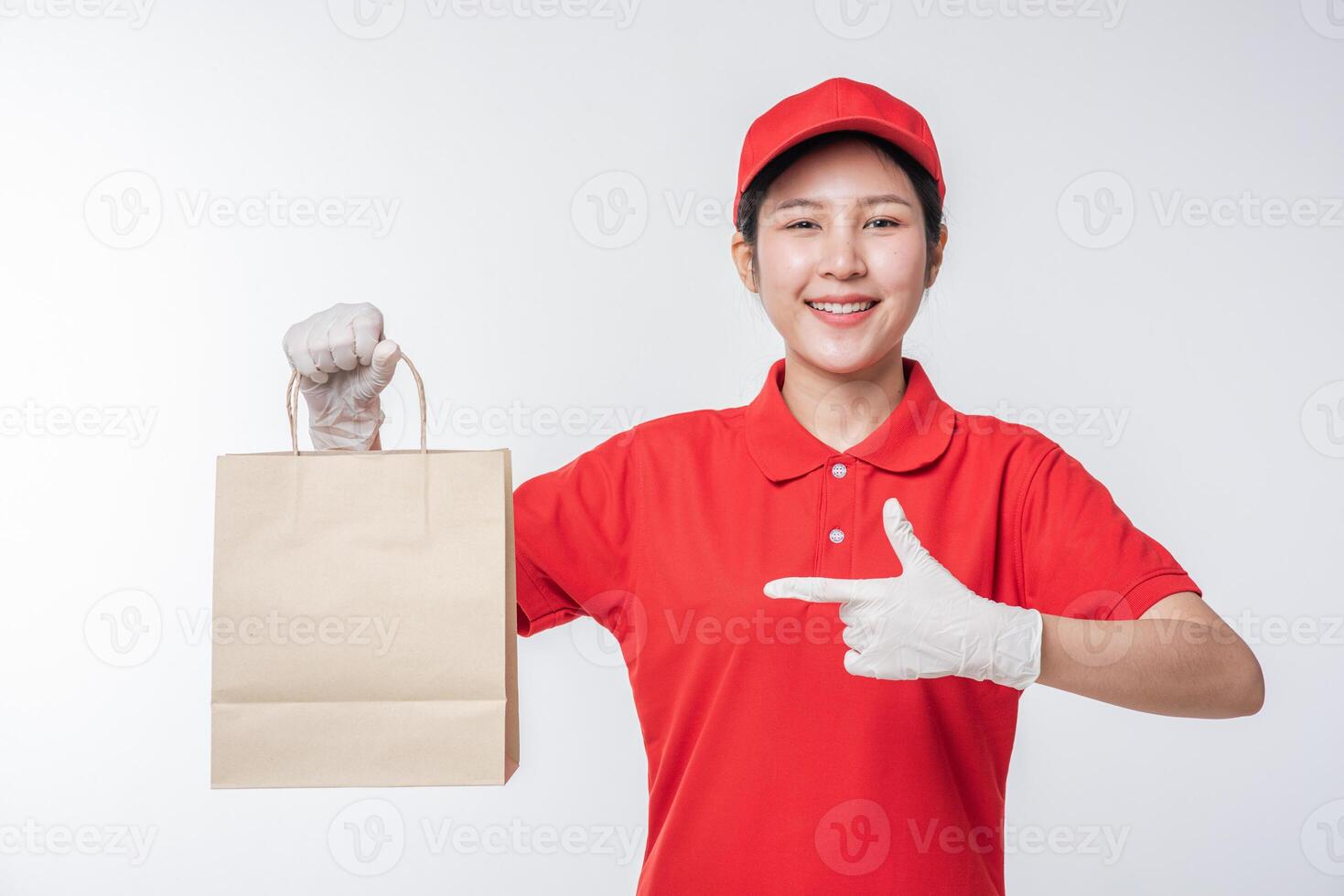 Image of a happy young delivery man in red cap blank t-shirt uniform standing with empty brown craft paper packet isolated on light gray background studio photo