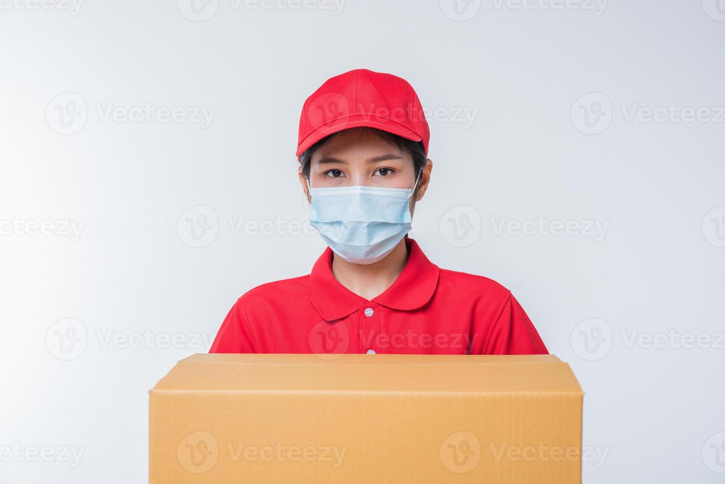 imagen de un joven repartidor consciente con gorra roja en blanco camiseta uniforme guantes de máscara facial de pie con una caja de cartón marrón vacía aislada en un estudio de fondo gris claro foto