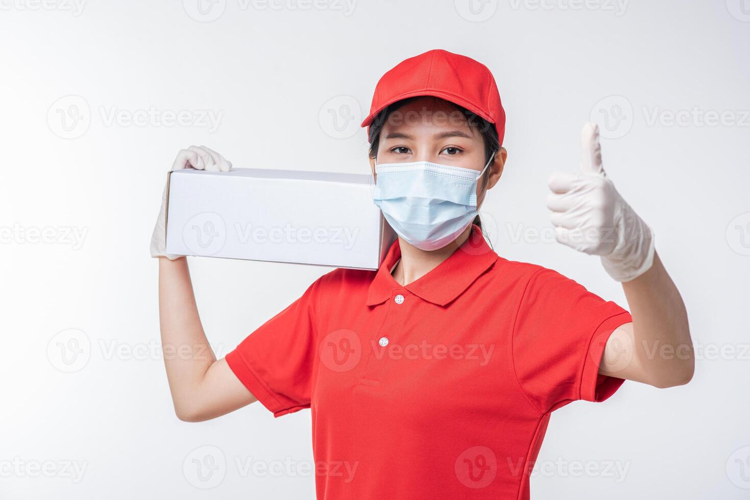 Image of a conscious young delivery man in red cap blank t-shirt uniform face mask gloves standing with empty white cardboard box isolated on light gray background studio photo