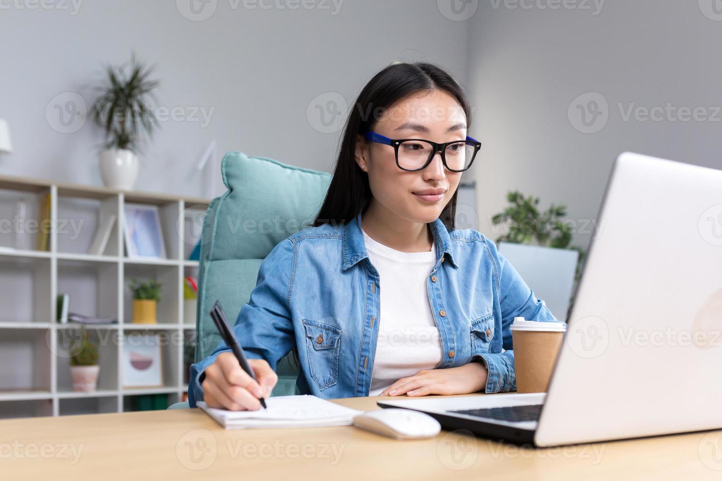 Young Asian woman studying online while sitting in office, writing lecture notes in notebook photo