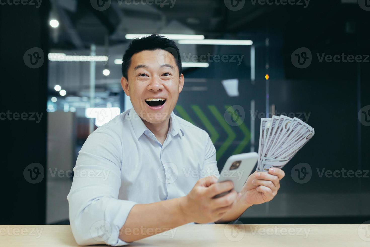 A happy young Asian man holds a telephone and cash money in his hands in office photo