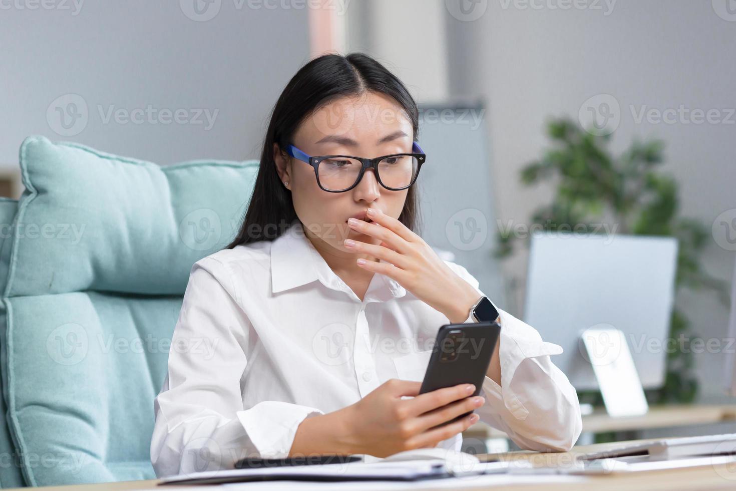 preocupado joven asiático negocio mujer en lentes sostiene el teléfono en su manos, sorprendido, conmocionado. foto