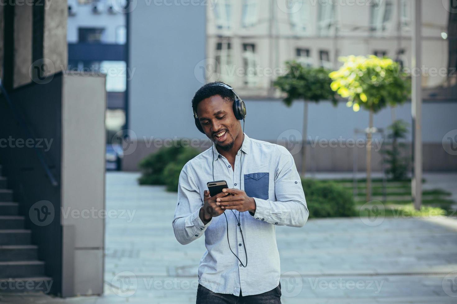 hombre escuchando a música usos grande auriculares, camina abajo el calle, contento africano americano foto