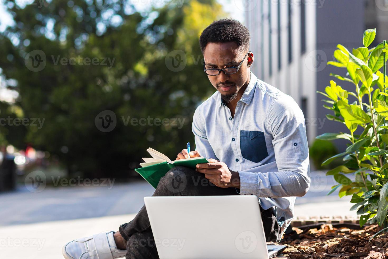 Young african american male student sitting in a city park on a bench with laptop and notebook photo