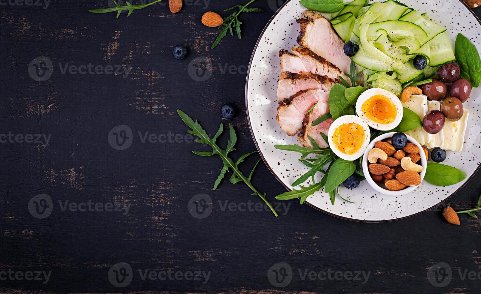 Ketogenic diet. Keto brunch. Boiled egg, pork steak and olives, cucumber, spinach, brie cheese, nuts and blueberry. Top view photo