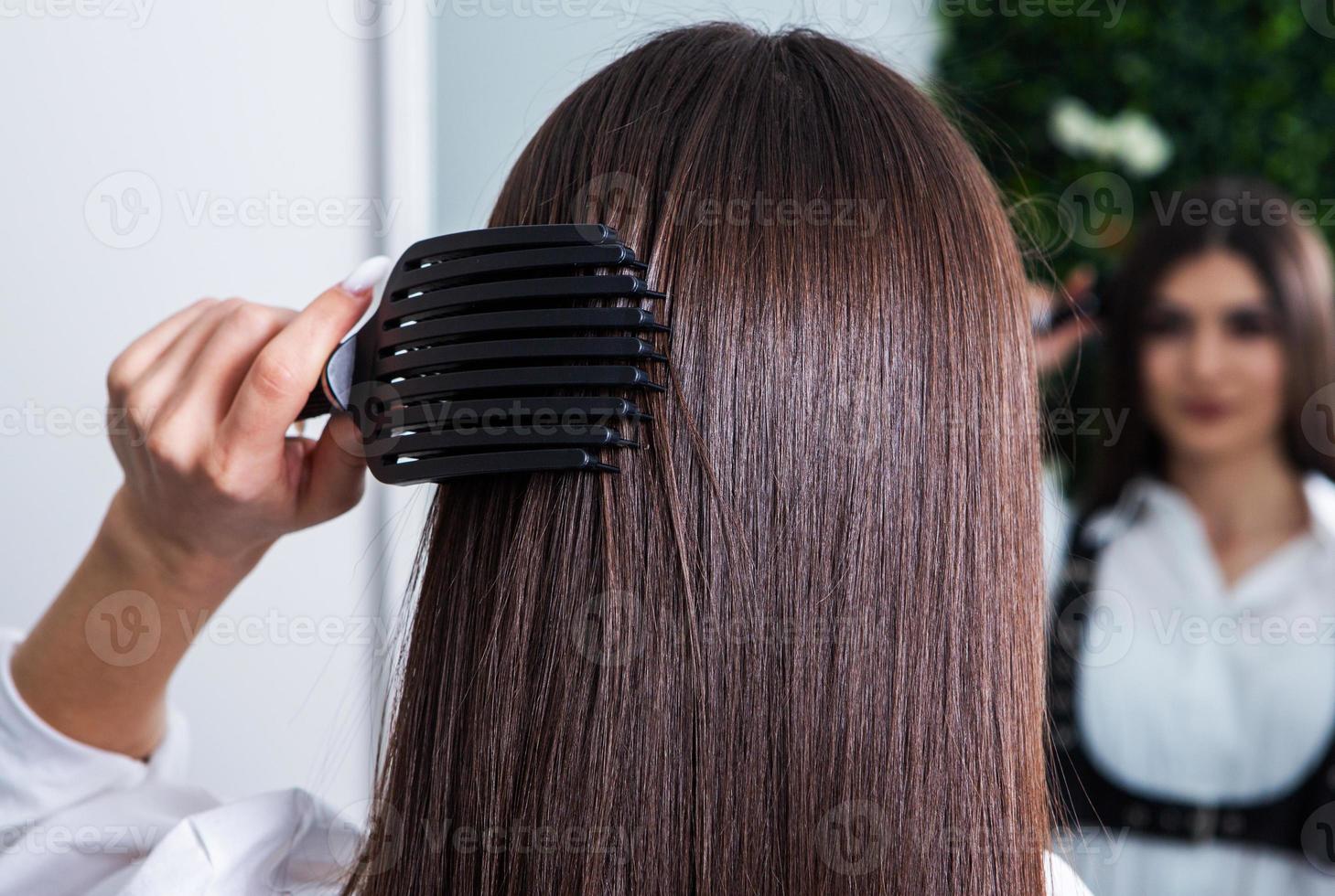 Young woman combing her long dark hair with a comb in a beauty salon. A straight healthy brunette hair that has undergone the hair straightening procedure. photo