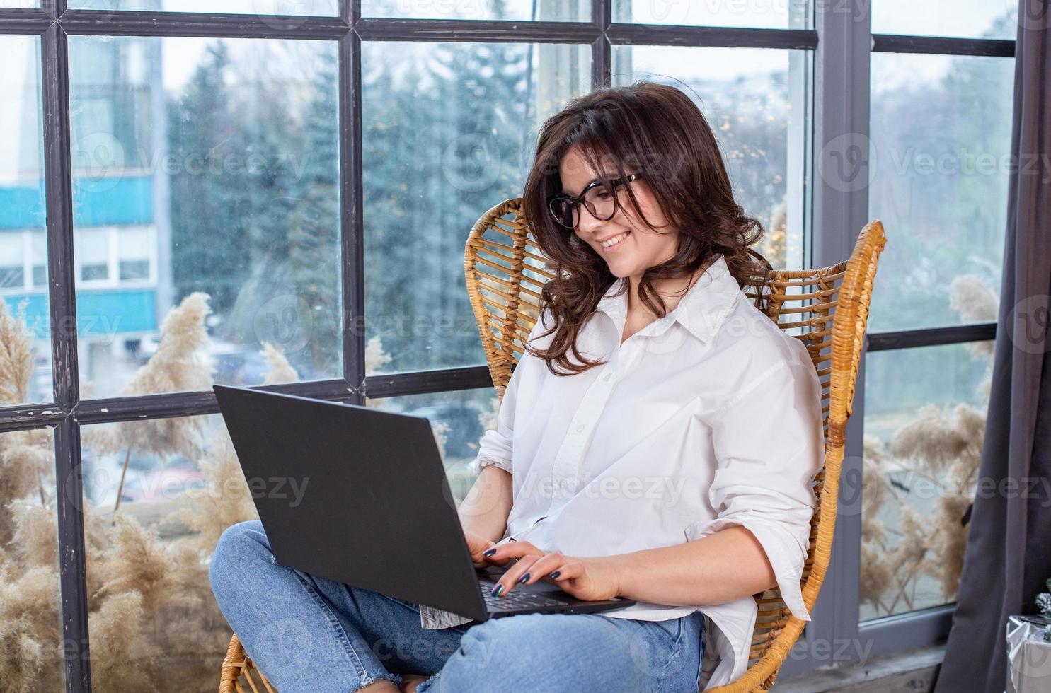 Beautiful businesswoman working on a laptop in a chair near the window. Female freelancer connecting to internet via computer. Blogger or journalist writing new article. photo