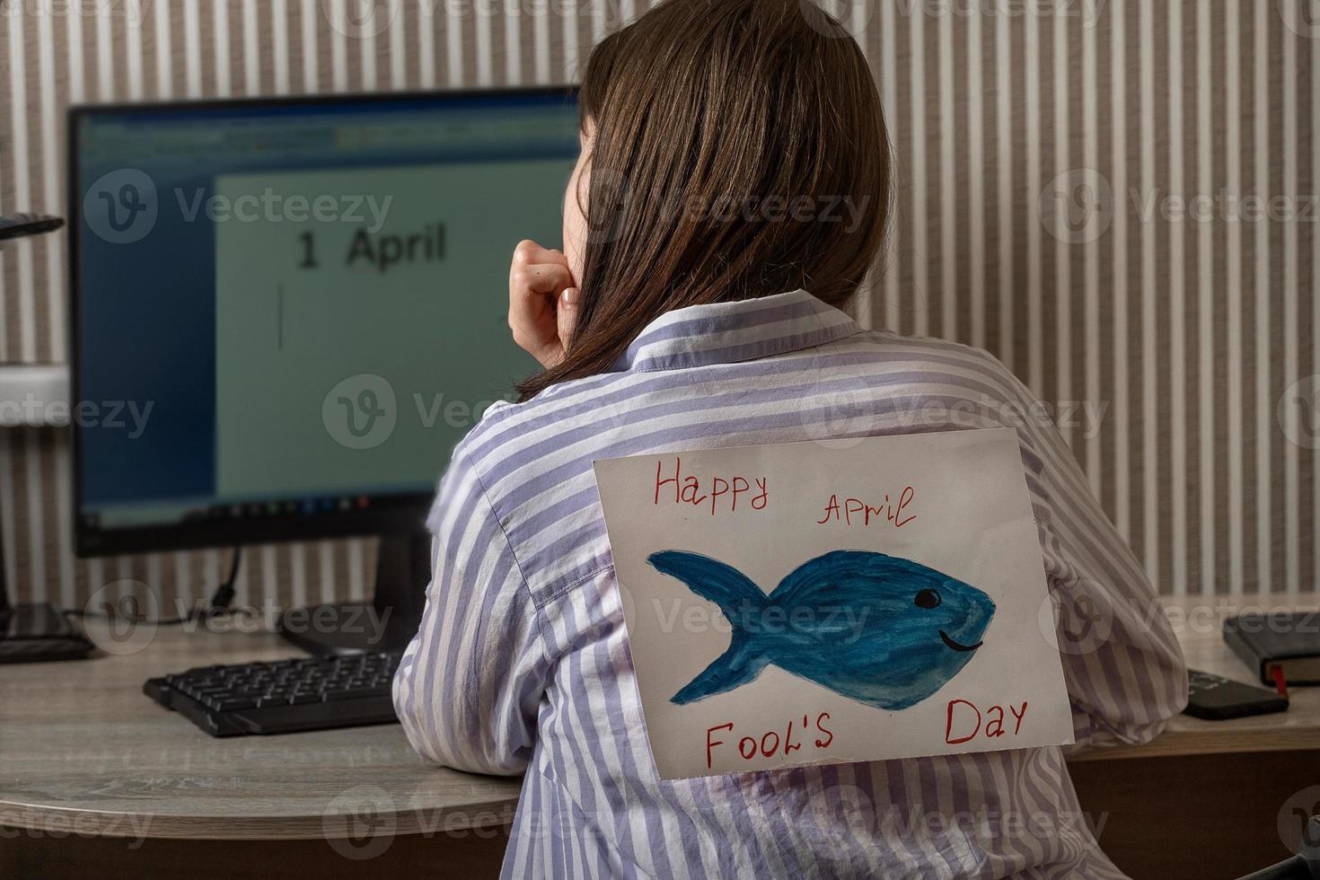A young woman with a paper fish from behind indoors works at a computer on which the inscription the first of April, April Fool's Day, jokes and jokes in the office on top of each other photo