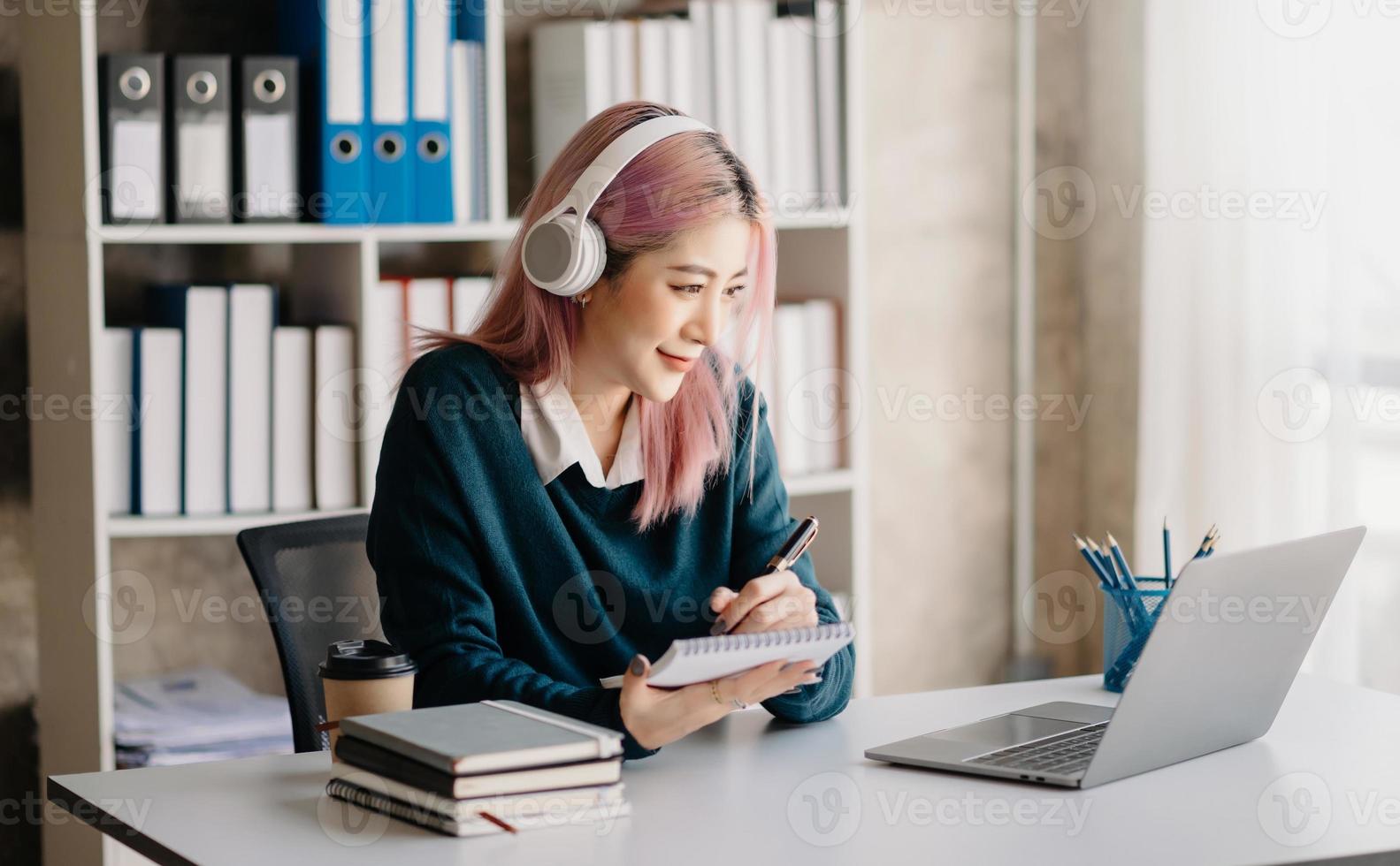 atractivo contento joven asiático estudiante estudiando a el Universidad biblioteca, sentado a el escritorio, utilizando un computadora portátil, tableta y auriculares teniendo un vídeo charlar. foto