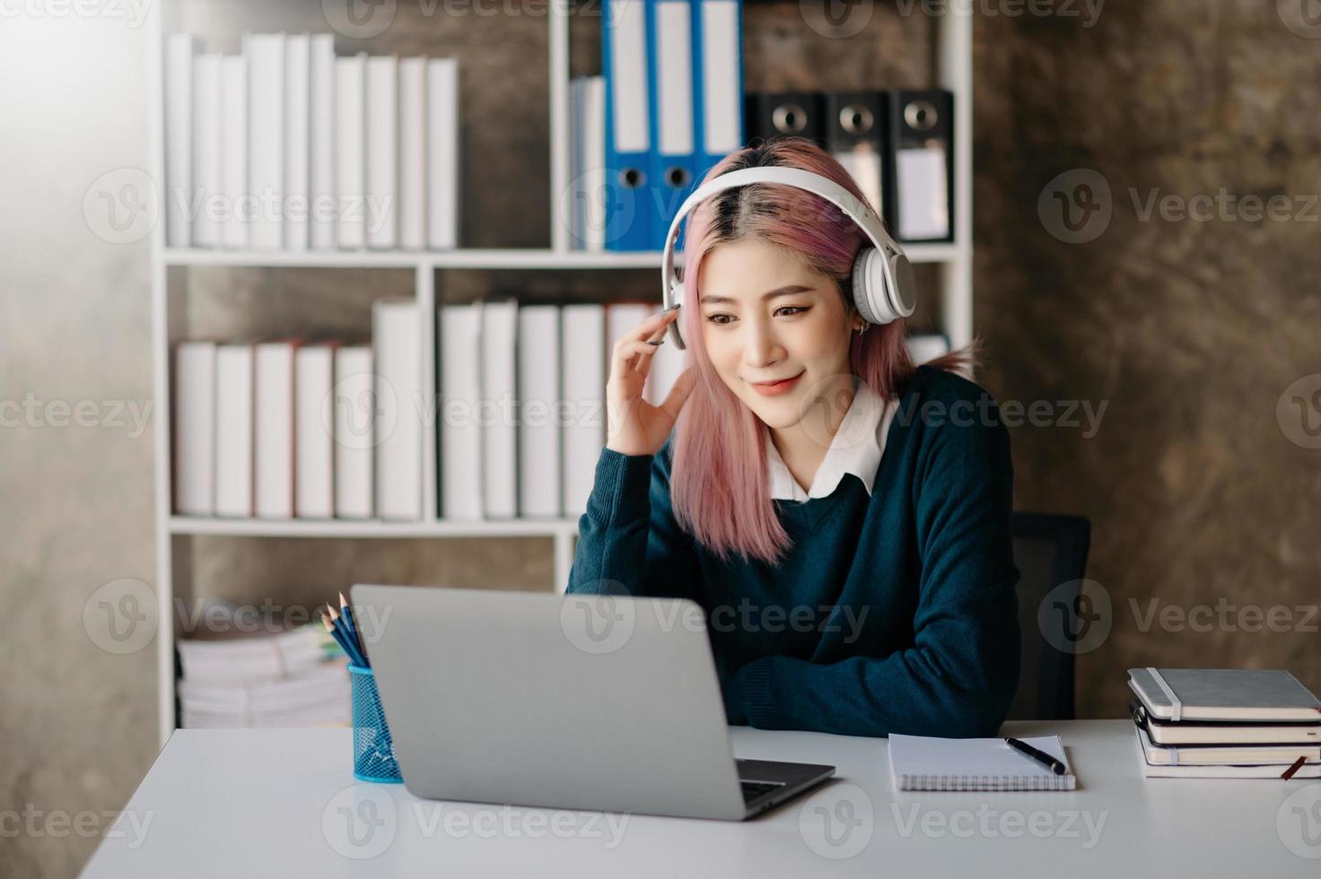 atractivo contento joven asiático estudiante estudiando a el Universidad biblioteca, sentado a el escritorio, utilizando un computadora portátil, tableta y auriculares teniendo un vídeo charlar. foto