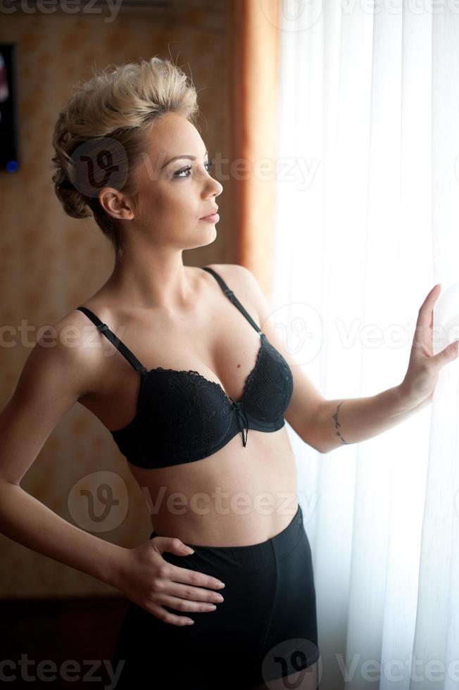 Attractive fair hair model with pantyhose and black bra and long gloves  posing provocatively. Fashion portrait of sensual blonde, studio shot.  Sensual female in black lingerie posing against window 20069703 Stock Photo