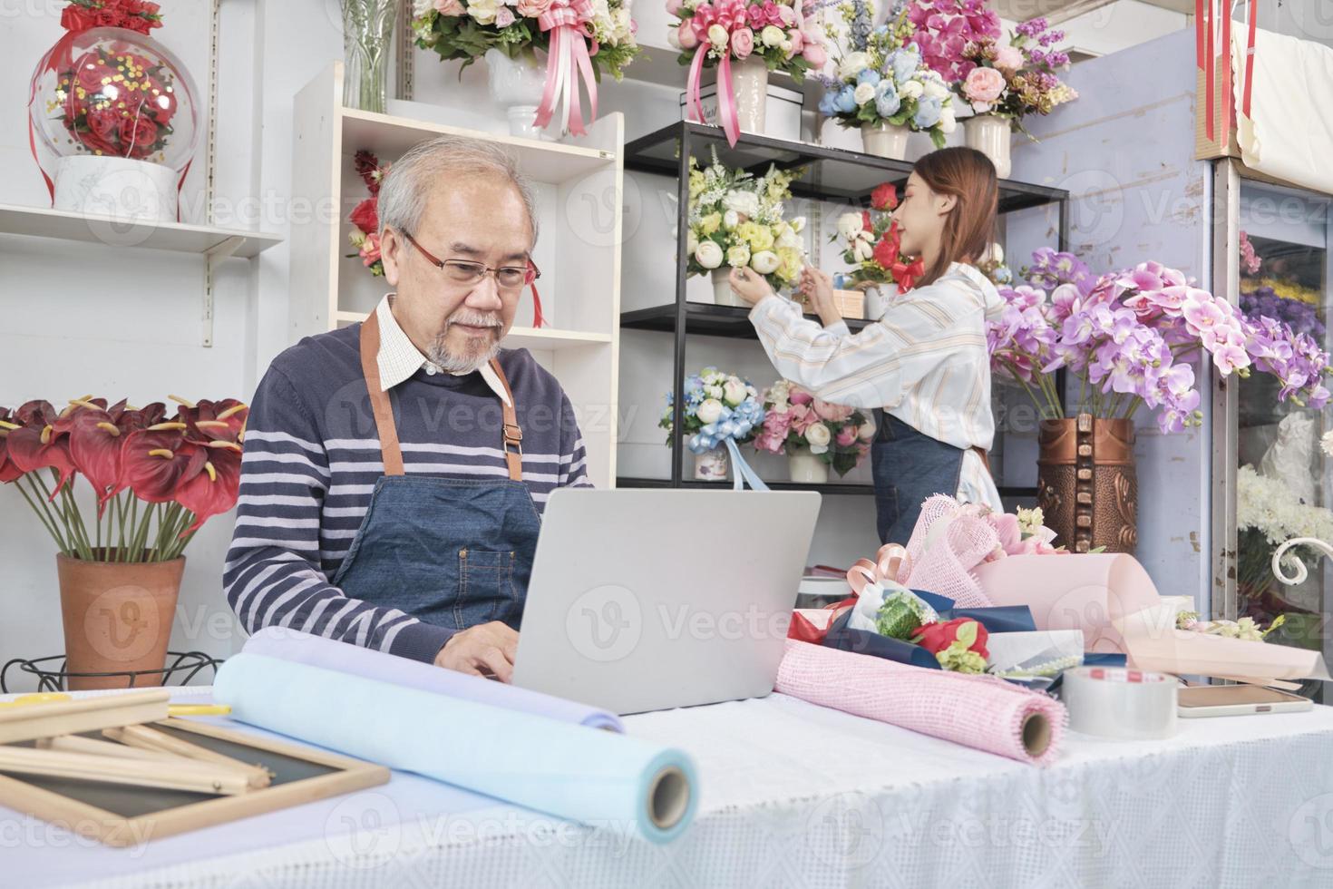 el anciano propietario asiático de una floristería que trabaja con una laptop en un sitio web para negocios en línea, una joven y hermosa comerciante que organiza flores frescas detrás, una colorida tienda de flores, una pyme de comercio electrónico. foto