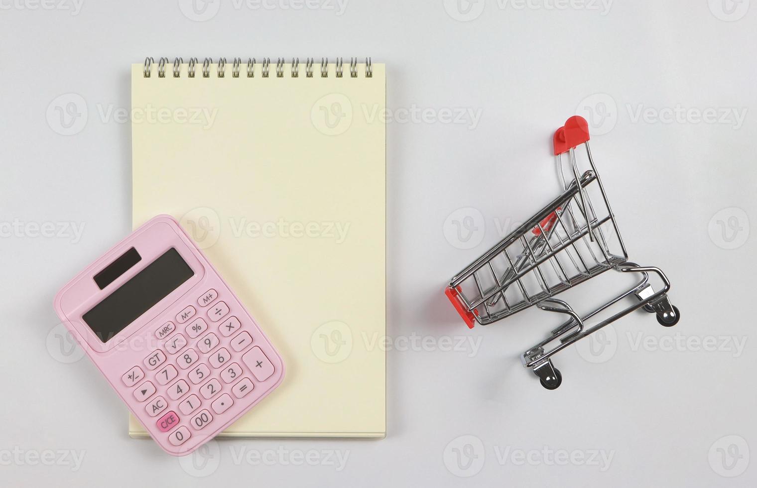 flat lay of pink calculator on blank page opened notebook and trolley or shopping cart on white background.. shopping budget, business and education concept. photo