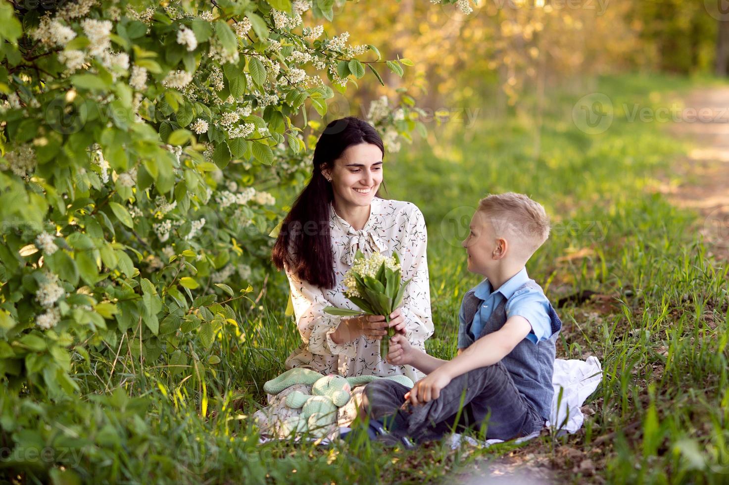 Mom is sitting next to her son on the grass , the boy is holding flowers in his hands photo