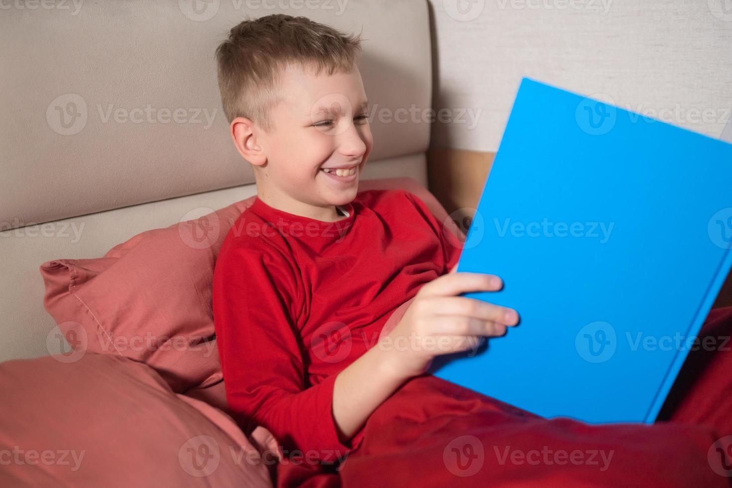 un contento chico en rojo pijama es acostado en cama y leyendo un libro foto