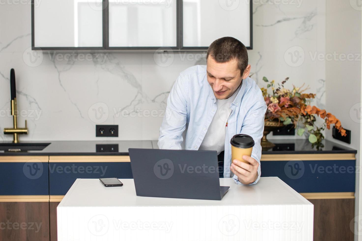 A young man standing in the kitchen is working on a laptop and holding a disposable cup photo