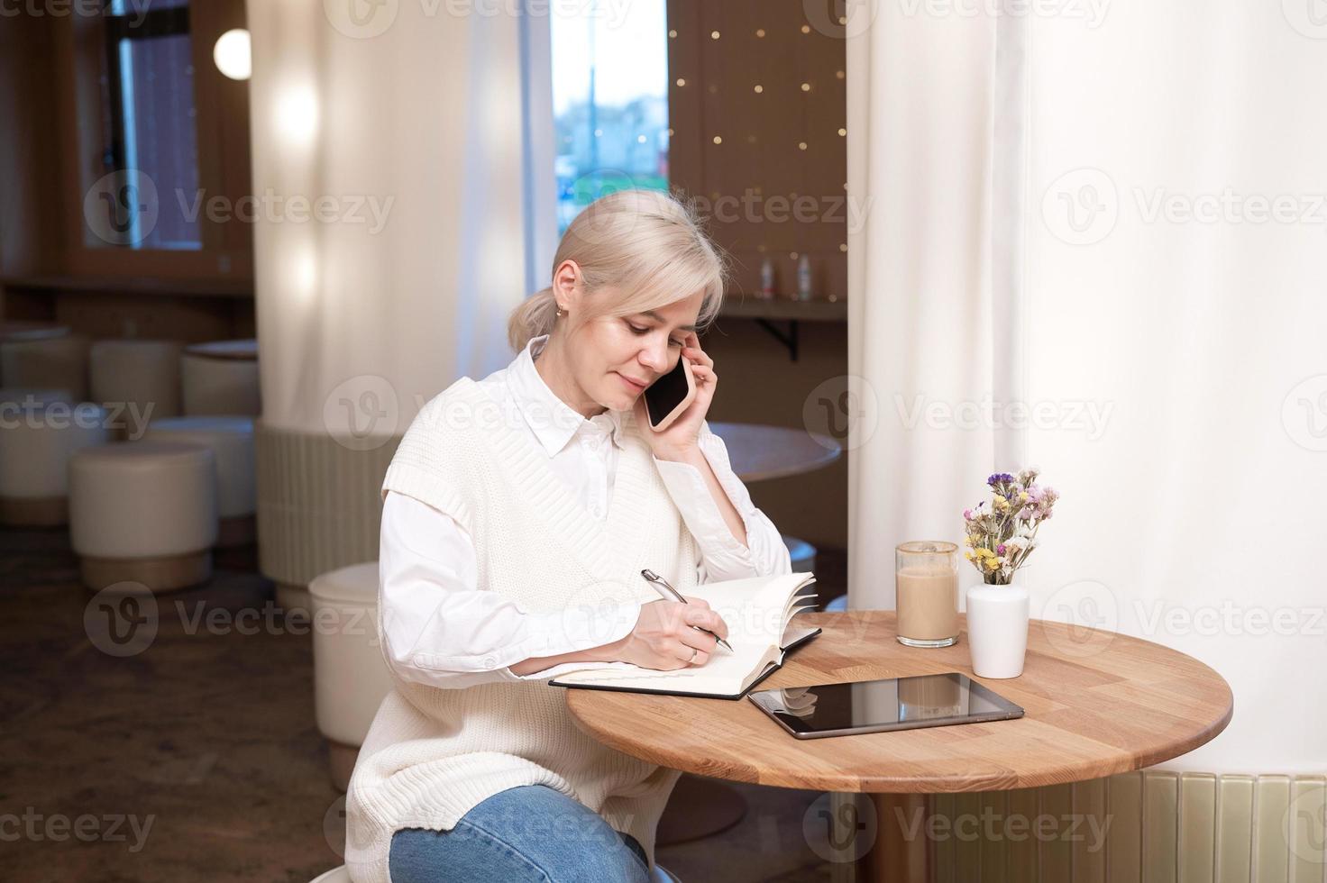 A cute girl is sitting in a cafe and talking on the phone and making notes in a diary photo