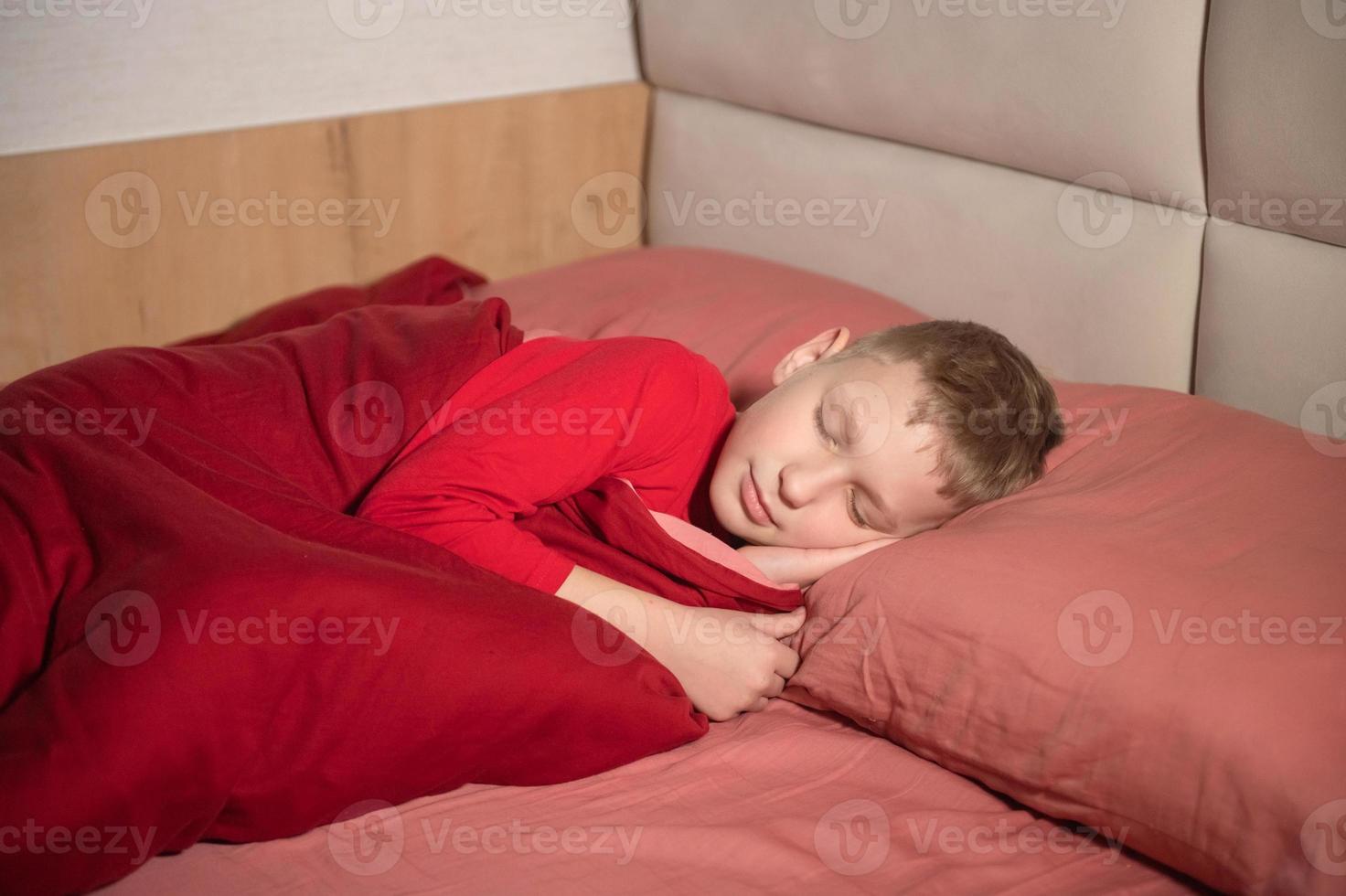 A cute boy in red pajamas is lying on a pillow with his eyes closed in bed and smiling photo