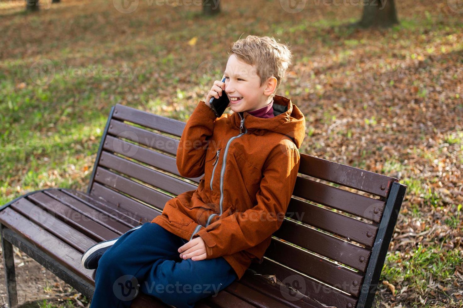 un chico es hablando en el teléfono y riendo mientras sentado en un parque banco en otoño foto