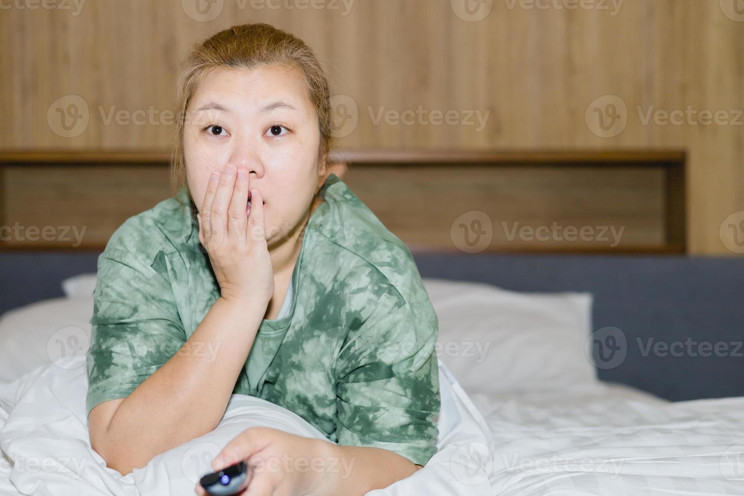 Asian housewife lying prone and look shocked in bed and holding remote control watching television with relaxing action. Hobby and free time life style concept. photo