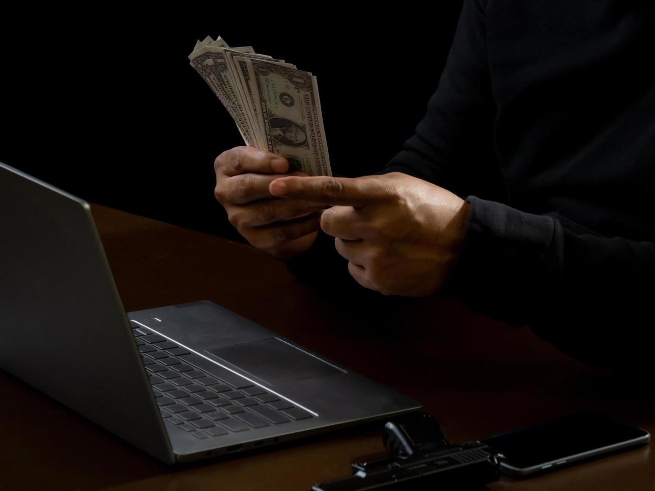 computadora y manos de un hombre con una camisa negra, sentado en una silla y una mesa, es un ladrón, sosteniendo dinero, contando la cantidad obtenida del secuestro o robo, en una habitación a oscuras. foto