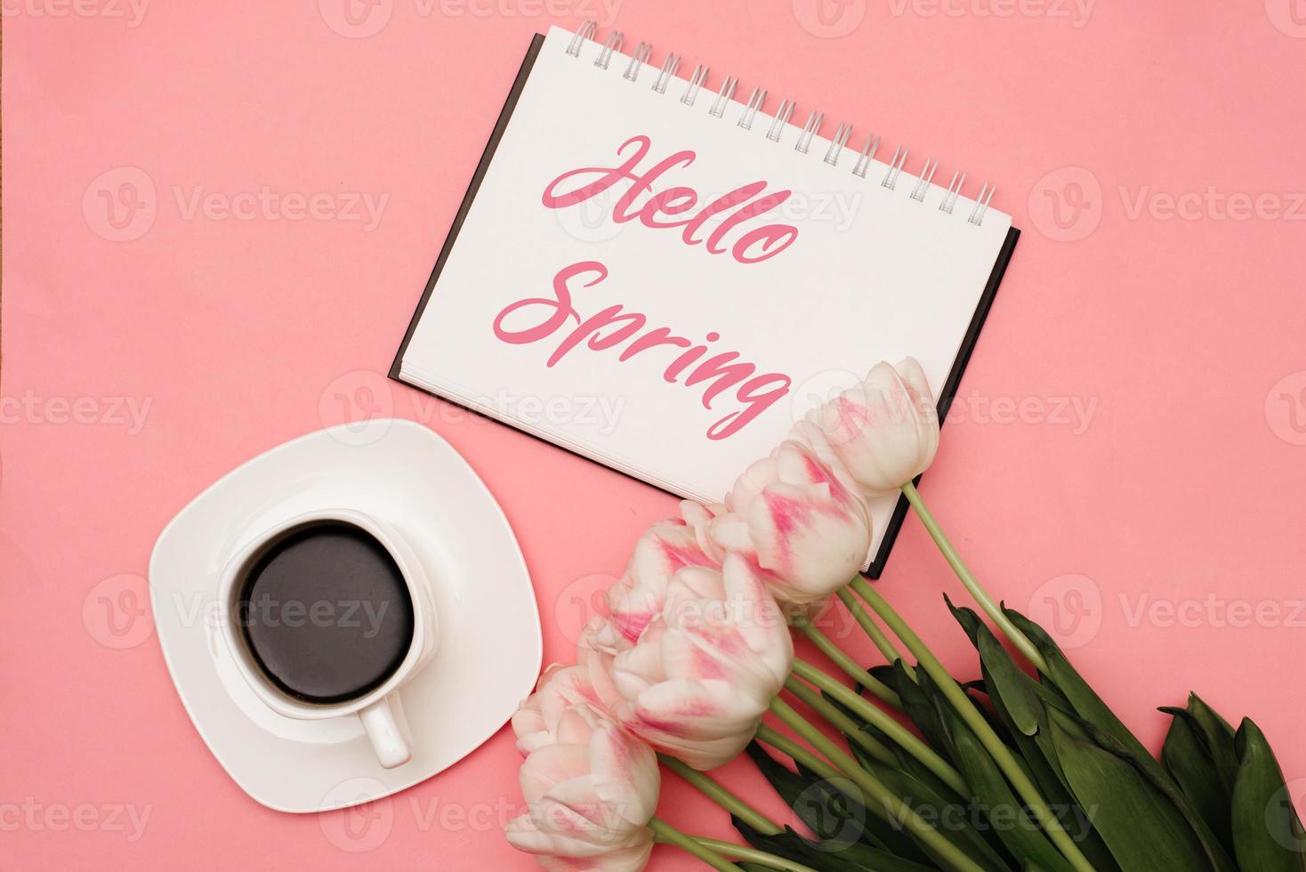 Text HELLO SPRING next to a beautiful bouquet of delicate pink tulips and a cup of coffee on a pink background. There may be your text. photo
