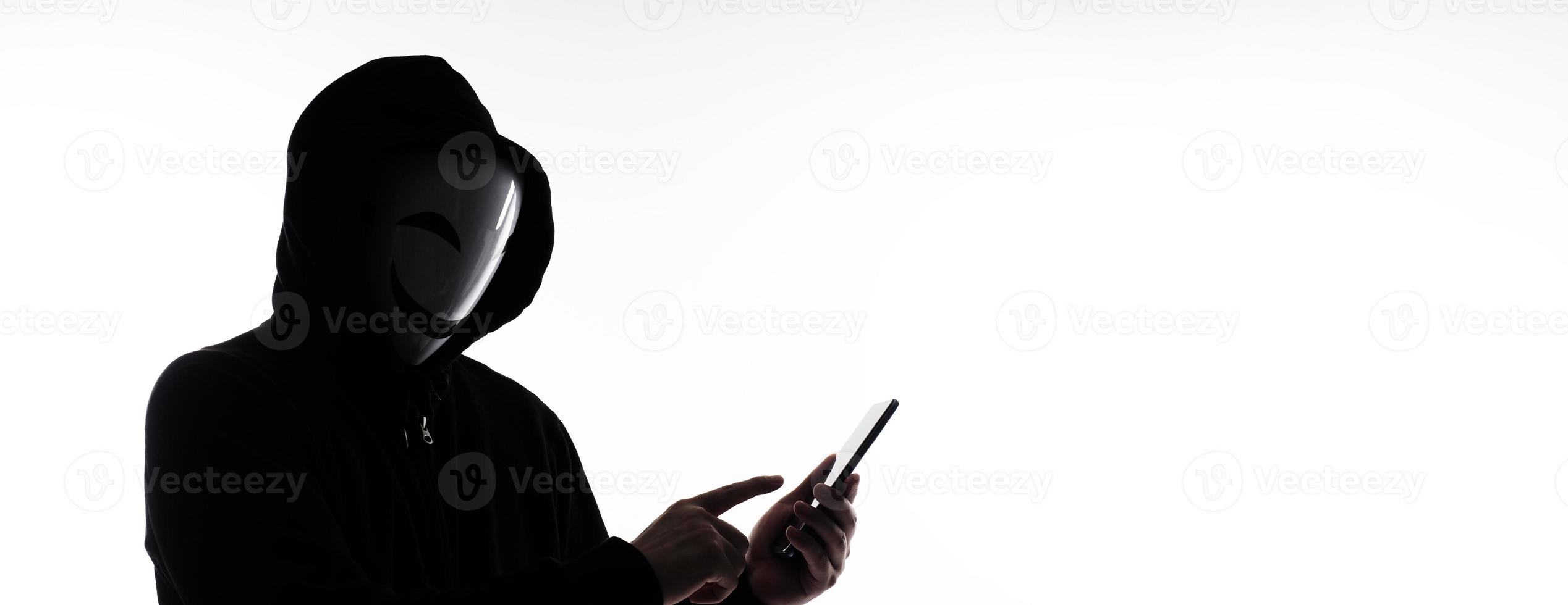 Hacker Anonymous and face mask with smartphone in hand. Man in black hood shirt holding and using mobile phone on white background. Represent cyber crime data hacking or stealing personal data concept photo