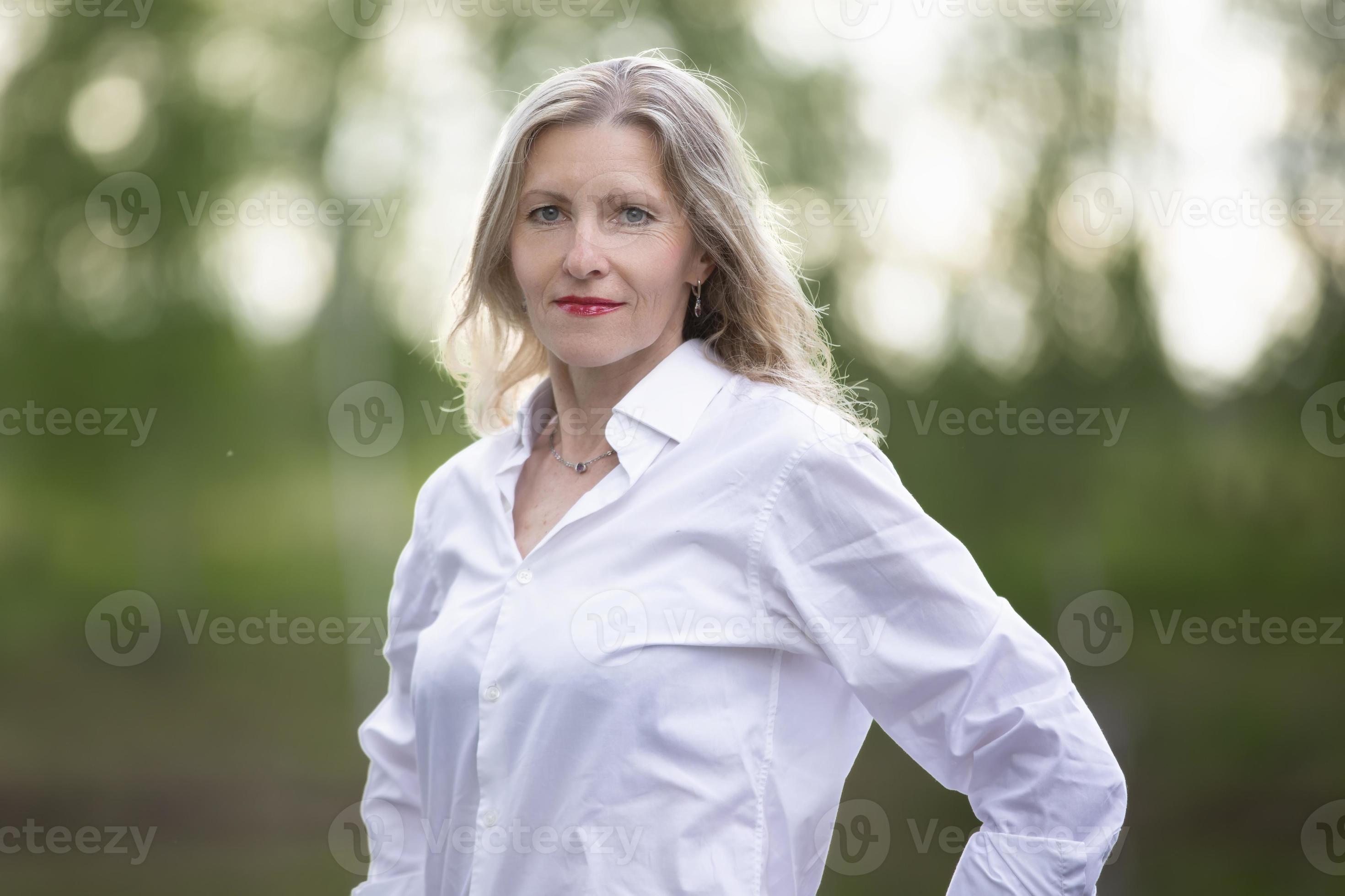 A beautiful middle-aged European woman. Fifty-year-old blonde woman in a  white shirt on the background of nature. 20129248 Stock Photo at Vecteezy