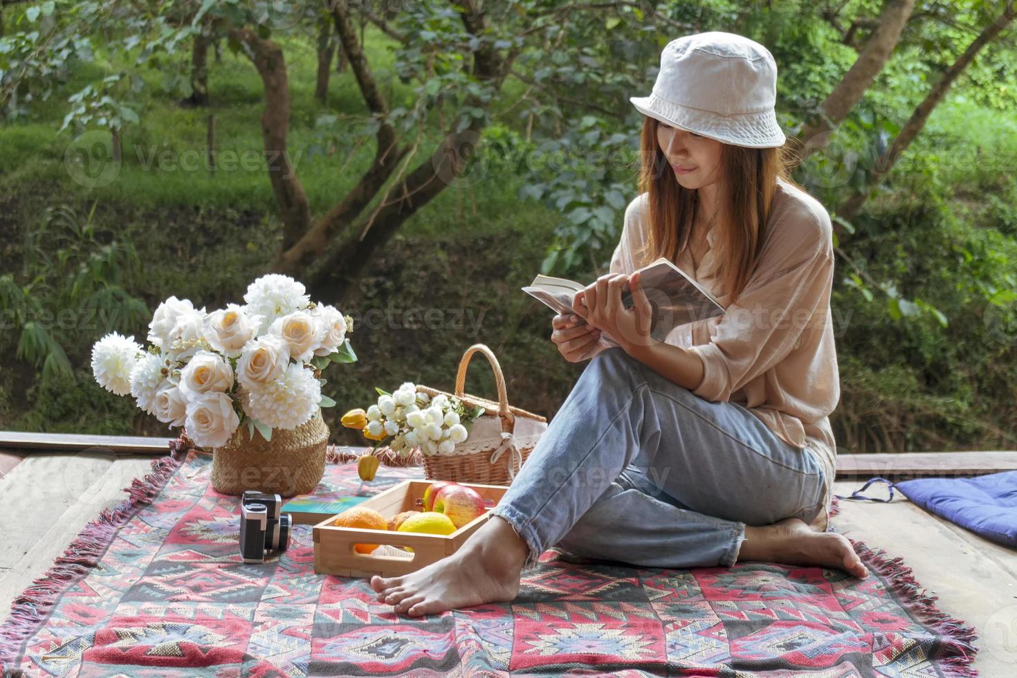 Woman in hat and old-fashioned dress sits and reads a book on a wooden balcony against a green forest backdrop. photo