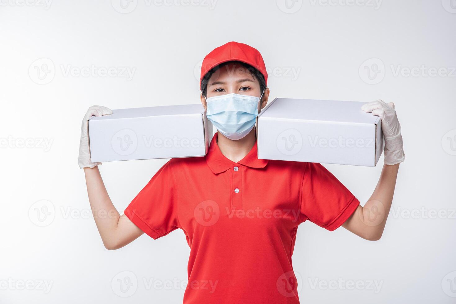 Image of a conscious young delivery man in red cap blank t-shirt uniform face mask gloves standing with empty white cardboard box isolated on light gray background studio photo