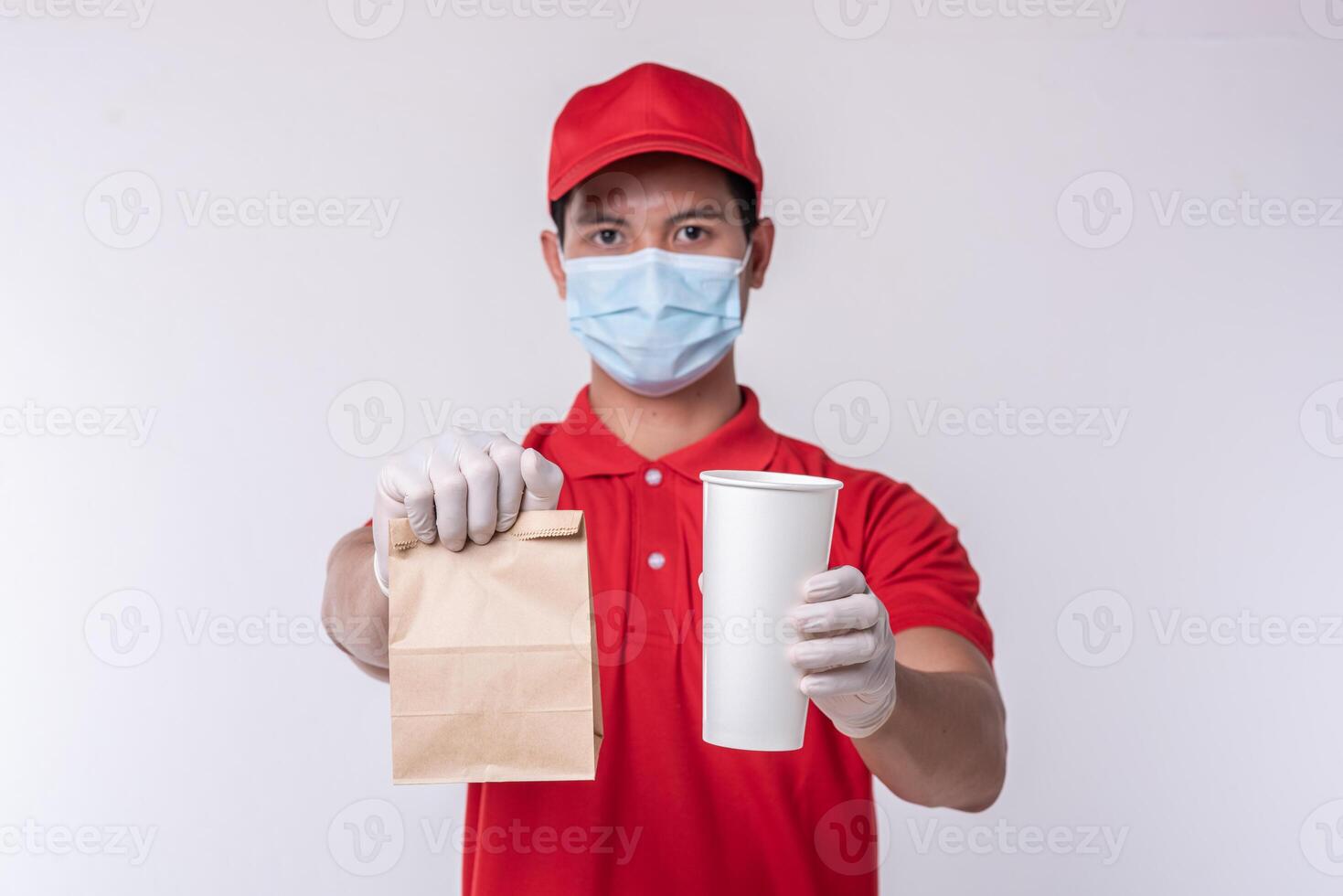 Image of a happy young delivery man in red cap blank t-shirt uniform face mask gloves standing with empty brown craft paper packet isolated on light gray background studio photo