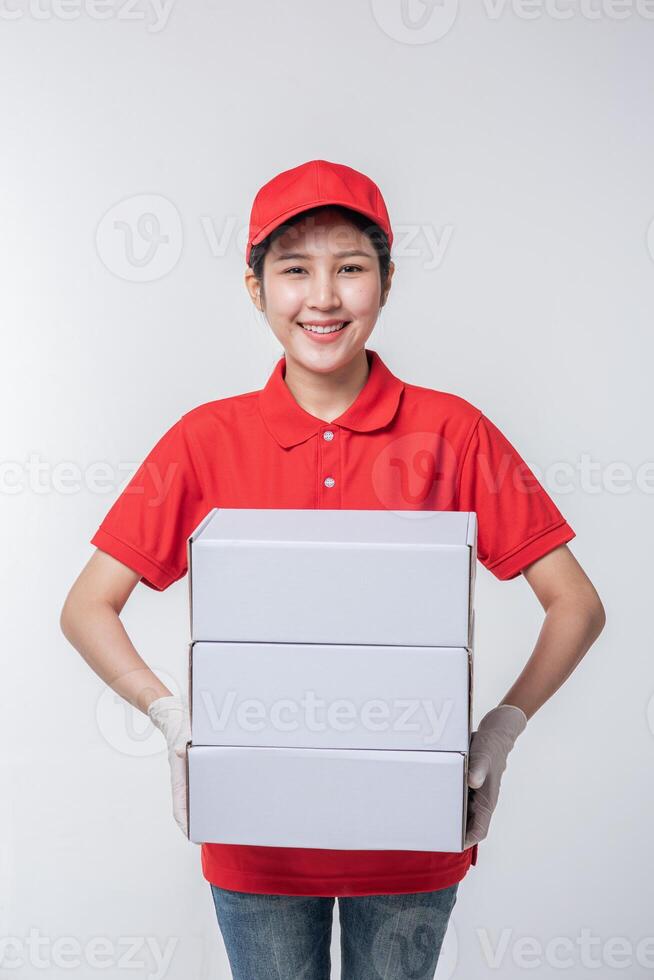 imagen de un joven repartidor con uniforme de camiseta en blanco con gorra roja de pie con una caja de cartón blanca vacía aislada en un estudio de fondo gris claro foto