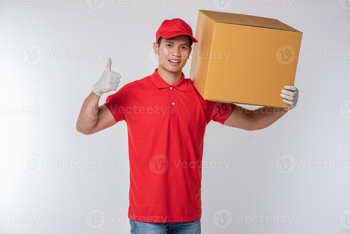 imagen de un joven y feliz repartidor con gorra roja, camiseta en blanco, uniforme de pie con una caja de cartón marrón vacía aislada en un estudio de fondo gris claro foto