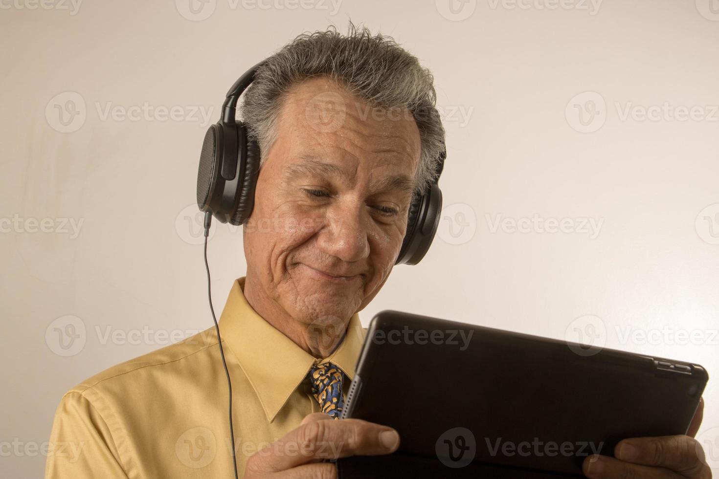 Older Man listening to music or a podcast wearing head phone on his smart tablet photo