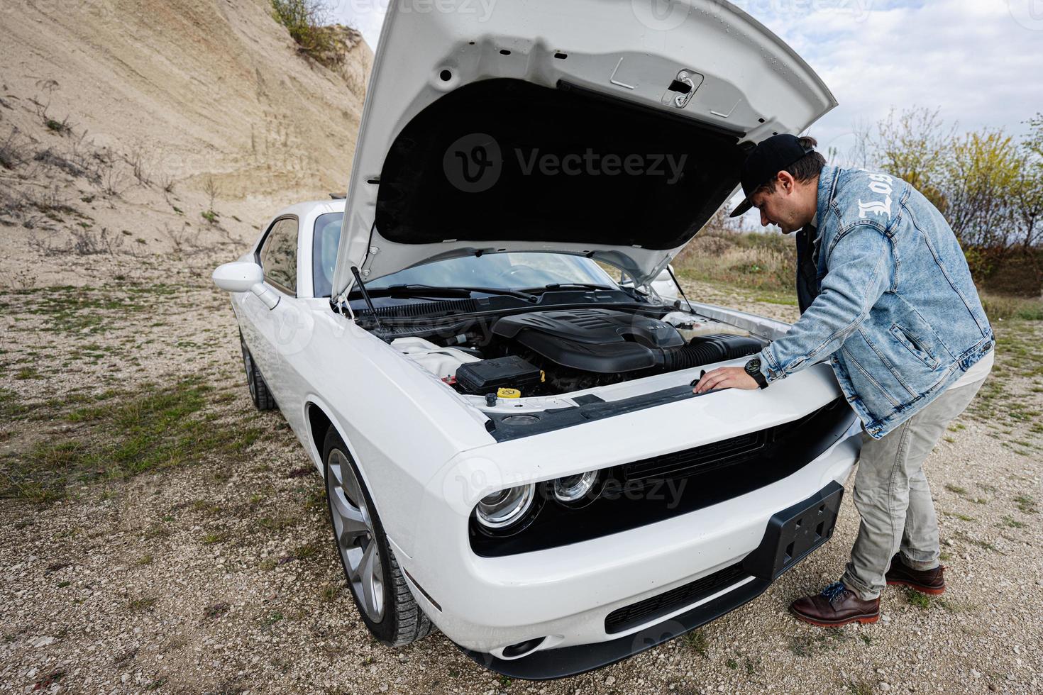 un hombre guapo con chaqueta de jeans y gorra está parado cerca de su auto blanco con capó abierto, revisa el motor. foto